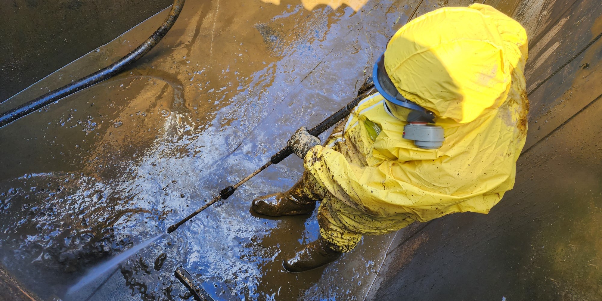 NEDT technician in protective suit and mask pressure washing industrial property.