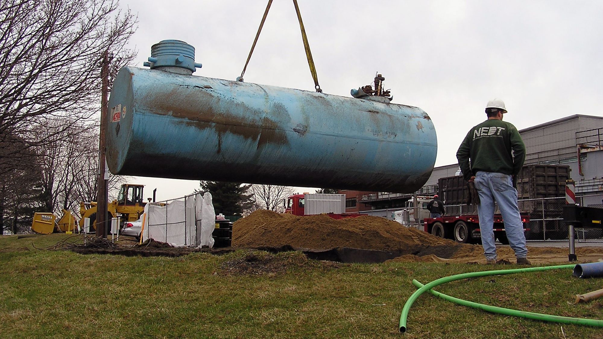 Crane lifts underground storage tank being supervised by NEDT engineer.